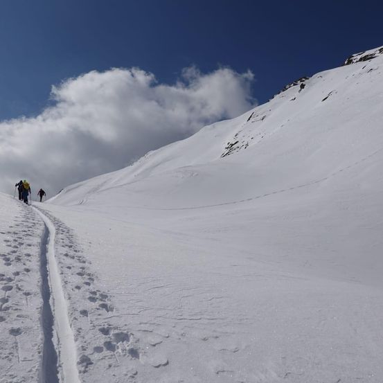 Einige Personen wandern auf einem verschneiten Berg unter klarem Himmel. Fußabdrücke sind im Schnee sichtbar.