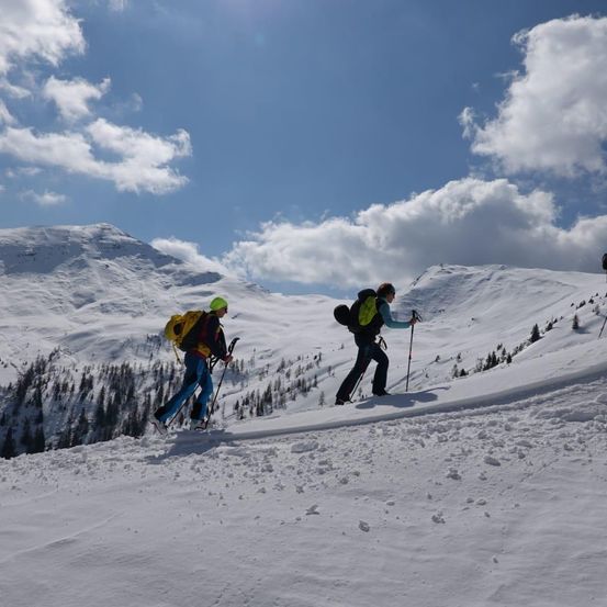 Zwei Personen sind im Schneegebirge Skifahren. Sie tragen gelbe Jacken, Helme und Rucksäcke. Sie halten Skistöcke in den Händen.