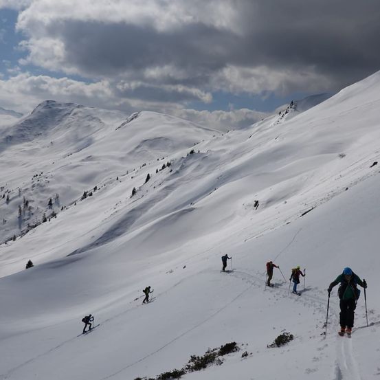 Eine Gruppe von Skifahrern erklimmt einen schneebedeckten Berghang. Sie benutzen Skistöcke, um aufzusteigen, mit einem bewölkten Himmel und entfernten Bergen im Hintergrund.