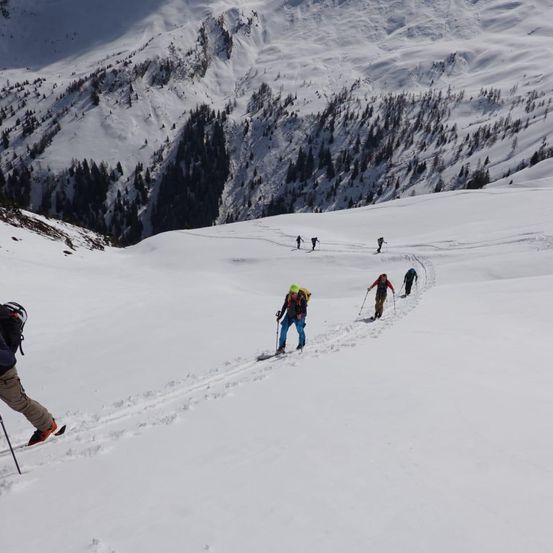 Fünf Skifahrer durchqueren einen verschneiten Berg. Sie sind mit Rucksäcken und Skistöcken ausgestattet. Das Gelände ist steil und schneebedeckt.