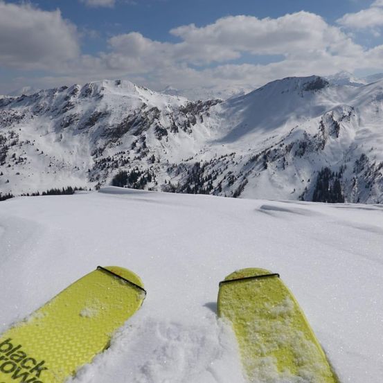 Schneebedeckte Berge aus der Perspektive eines Skifahrers. Heller, blauer Himmel mit Wolken. Gelbe Skier mit dem Wort 'Black Crow' sichtbar.