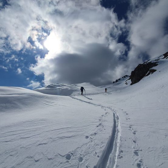 Zwei Personen skifahren auf einem schneebedeckten Berg unter einem bewölkten Himmel. Die Sonne scheint durch die Wolken.
