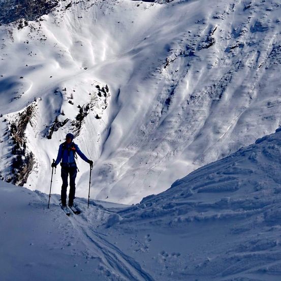 Ein Skifahrer in Blau fährt mit Skistöcken einen schneebedeckten Hang hinauf, unter einem hellen Sonnenschein.