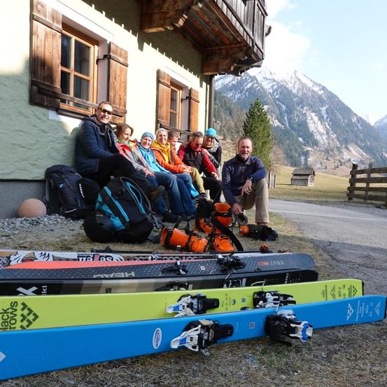 Mehrere Skifahrer posieren vor einem Chalet für ein Foto, mit Bergen im Hintergrund. Sie sitzen auf dem Boden mit ihren Skiern davor ausgebreitet.
