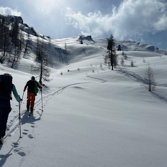 Zwei Personen treiben Langlauf auf einem verschneiten Berg. Einer trägt eine Mütze und beide haben Skistöcke. Bäume säumen die Landschaft.
