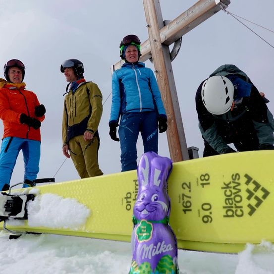 Vier Personen stehen im Schnee neben einem Skibrett. Ein riesiger Schokoladenhase liegt auf dem Brett. Sie sind alle in Winterkleidung und Helmen gekleidet.
