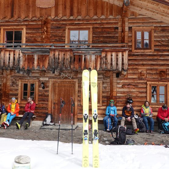 Skifahrer in Winterausrüstung sitzen um eine Holzhütte herum, Skier und Stöcke in den Schnee gestellt. Das Gebäude hat Holzfenster und eine Holztür. Der Schnee ist dick und weiß.