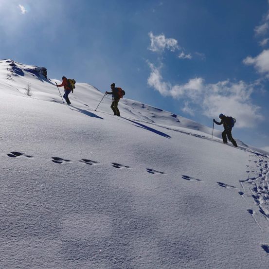 Drei Personen wandern einen verschneiten Berg hinauf. Sie tragen alle Rucksäcke und benutzen Skistöcke. Fußabdrücke sind im Schnee sichtbar.