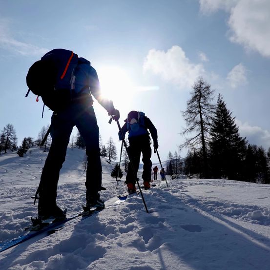 Zwei Skifahrer durchqueren einen verschneiten Berg, einer mit einem Rucksack und der andere mit Skistöcken. Die Sonne scheint hell und wirft Schatten auf den Schnee. Bäume und andere Skifahrer sind im Hintergrund sichtbar.