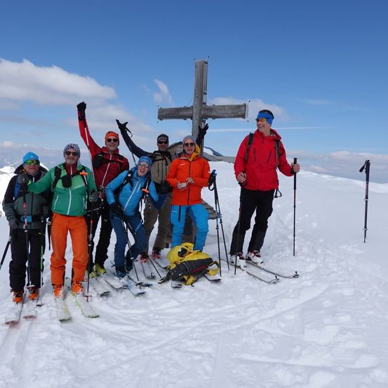 Eine Gruppe von Skifahrern posiert für ein Foto auf einem schneebedeckten Berggipfel mit einem Kreuz im Hintergrund.