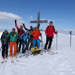 Eine Gruppe von Skifahrern posiert für ein Foto auf einem schneebedeckten Berggipfel mit einem Kreuz im Hintergrund.