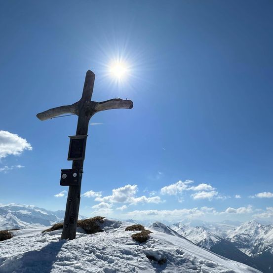 Ein holzkreuz steht auf einem verschneiten berggipfel unter einem hellen sonnenschein und blauem himmel mit einigen wolken.