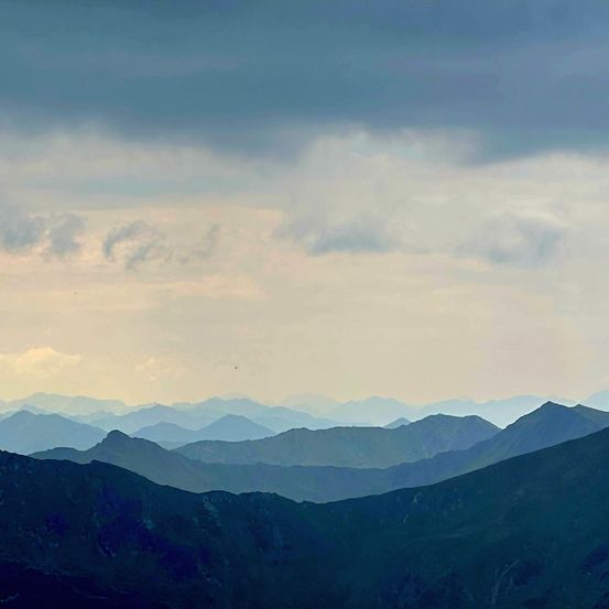 Ein Panoramablick auf eine Bergkette unter einem bewölkten Himmel. Die Berge sind gestaffelt, mit Blau- und Grautönen. Der Himmel ist bewölkt, mit Sonnenstrahlen, die durchbrechen.