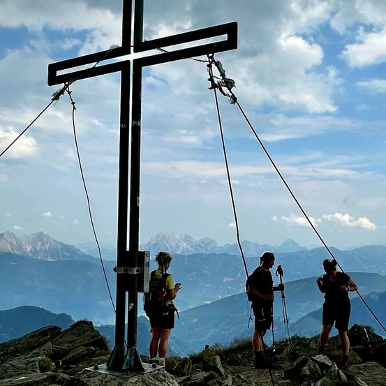 Drei Wanderer stehen auf einem Berggipfel mit einem großen Kreuz im Vordergrund, umgeben von felsigem Gelände und einer Kulisse aus majestätischen Bergen unter einem bewölkten Himmel.
