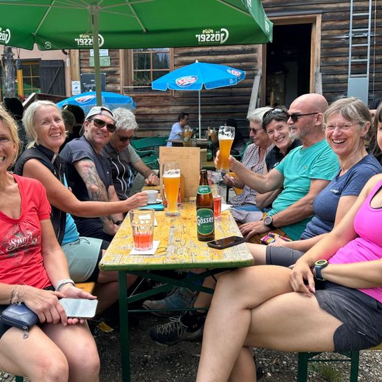 Eine Gruppe von Menschen sitzt um einen Tisch in einem Biergarten, lächelt und hält Gläser mit Bier. Über ihnen befinden sich zwei grüne Schirme.