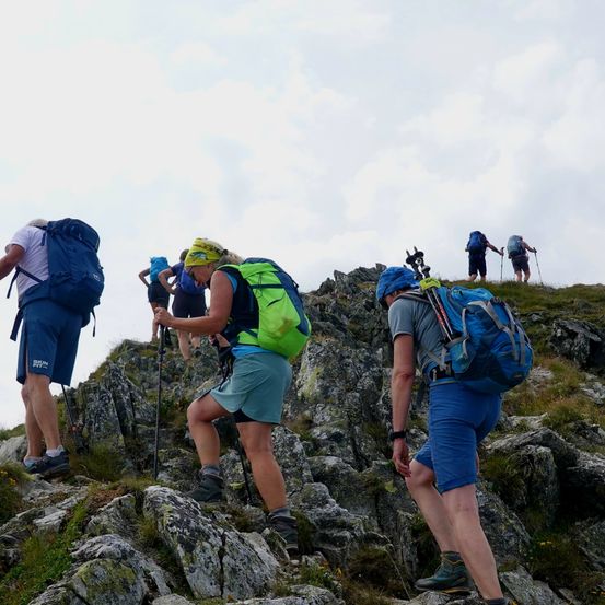 Eine Gruppe von Wanderern mit Rucksäcken klettert einen felsigen Bergpfad unter einem bewölkten Himmel hinauf.