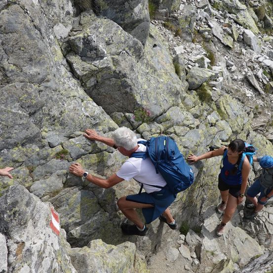 Drei Wanderer klettern einen felsigen Berghang hinauf. Der Mann vorne hat einen blauen Rucksack und hält sich an dem Felsen fest. Die Frau hinter ihm trägt ein blaues Tanktop und schwarze Shorts. Eine weitere Person ist rechts teilweise sichtbar.