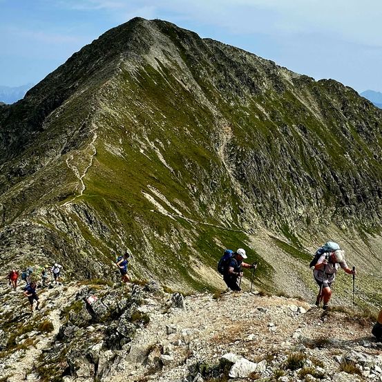 Eine Gruppe von Wanderern erklimmt einen steilen Bergpfad, wobei einige Trekkingstöcke zur Unterstützung benutzen. Der Weg ist schmal und steinig, mit Flecken von grünem Gras. Der Himmel ist klar und blau.