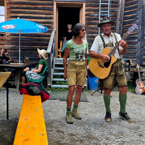 Ein Mann und eine Frau in traditioneller deutscher Kleidung spielen Gitarren vor einem Holzgebäude. Dahinter sitzt eine Frau auf einem Stuhl und ein Mann sitzt an einem Tisch unter einem Regenschirm.