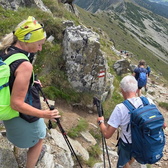 Eine Gruppe von Wanderern, einschließlich eines älteren Mannes und einer Frau, erklimmt einen felsigen Bergpfad. Sie sind mit Rucksäcken und Trekkingstöcken ausgestattet. Die Frau vorne trägt ein gelbes Kopftuch. In der Ferne sind andere Wanderer auf demselben Pfad zu sehen.
