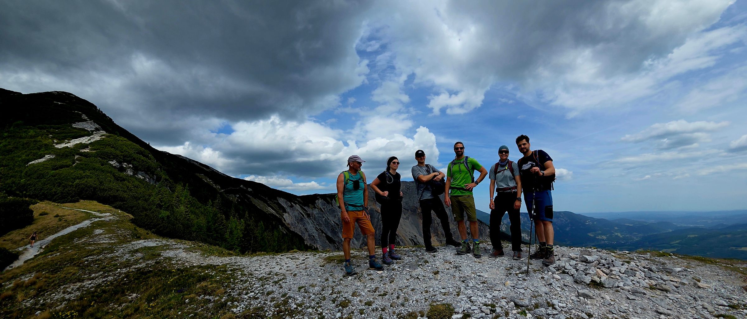 Sechs Menschen stehen auf einem felsigen Berggipfel, dem Betrachter zugewandt. Über ihnen hängen bewölkte Himmel. Sie sind alle zum Wandern gekleidet.