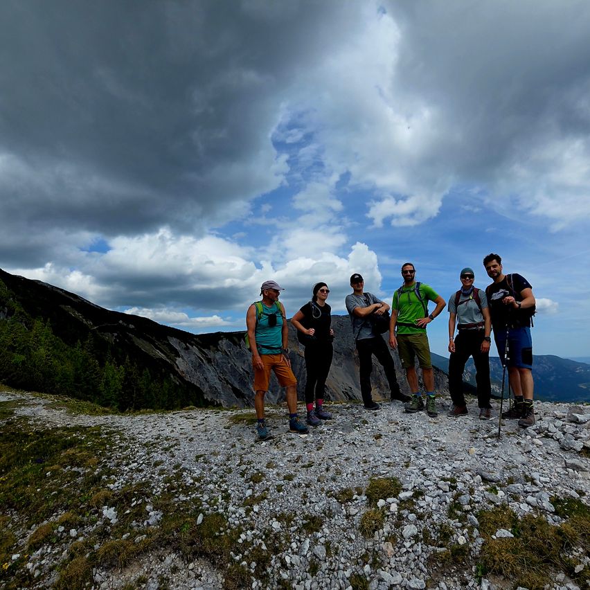 Sechs Wanderer, fünf Männer und eine Frau, posieren für ein Foto auf einem felsigen Gipfel unter einem teilweise bewölkten Himmel.