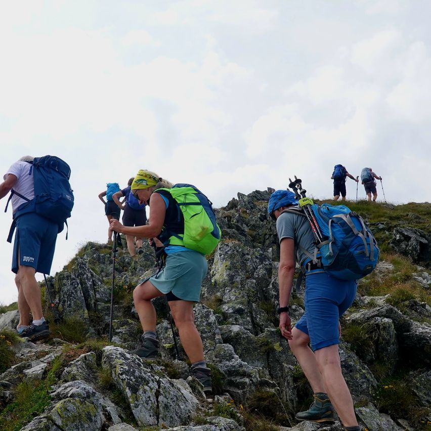 Eine Gruppe von Wanderern mit Rucksäcken klettert unter einem bewölkten Himmel einen felsigen Berg hinauf.