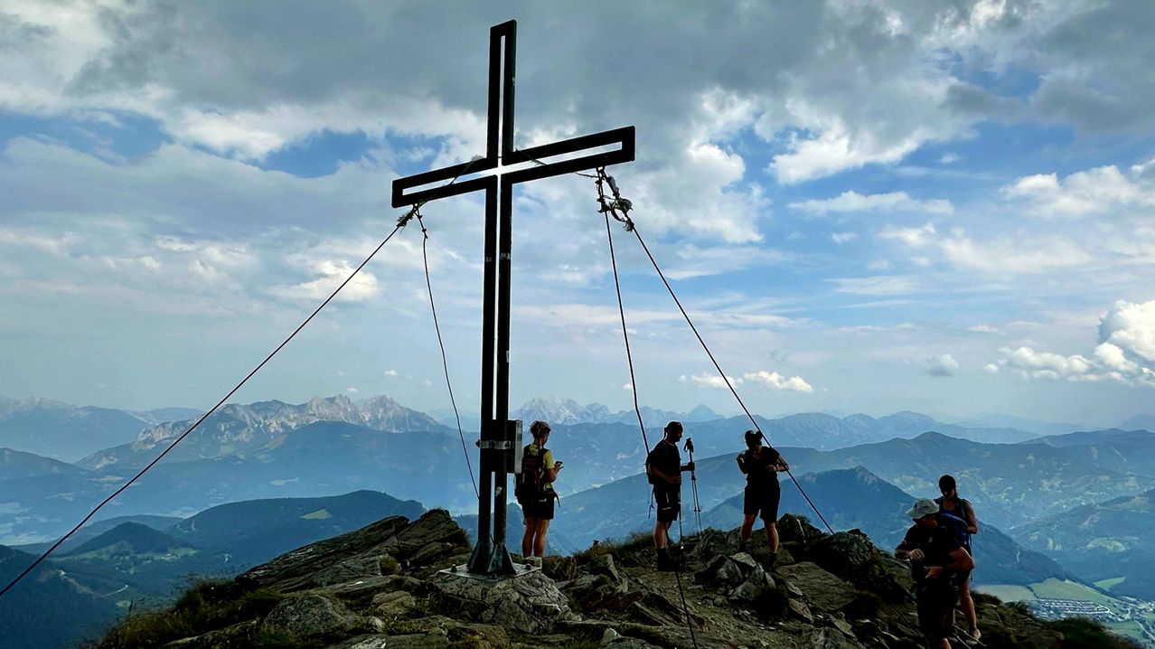 Drei Wanderer stehen auf einem Berggipfel mit einem großen Kreuz im Vordergrund, unter einem teilweise bewölkten Himmel, mit Blick auf die Berge im Hintergrund.