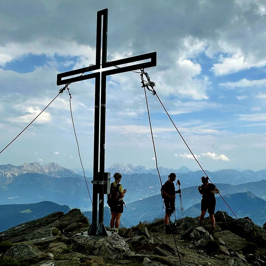 Drei Wanderer stehen auf einem Berggipfel mit einem großen Kreuz im Vordergrund, unter einem teilweise bewölkten Himmel, mit Blick auf die Berge im Hintergrund.