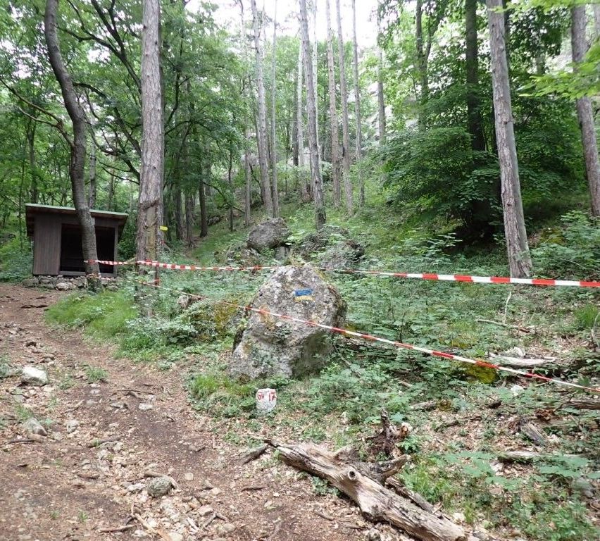 Ein hölzernes Gebäude steht in einem bewaldeten Gebiet, mit einem großen Felsen in der Mitte, umgeben von rot-weißem Warnschild.