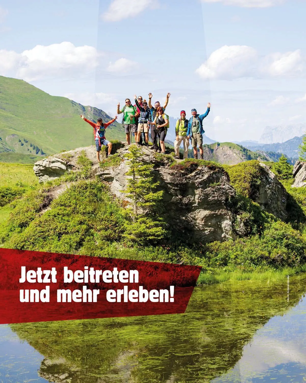 Eine Gruppe von Wanderern steht auf einem großen Felsen in einem Berggebiet, mit einem Grasfeld und einem Gewässer darunter.