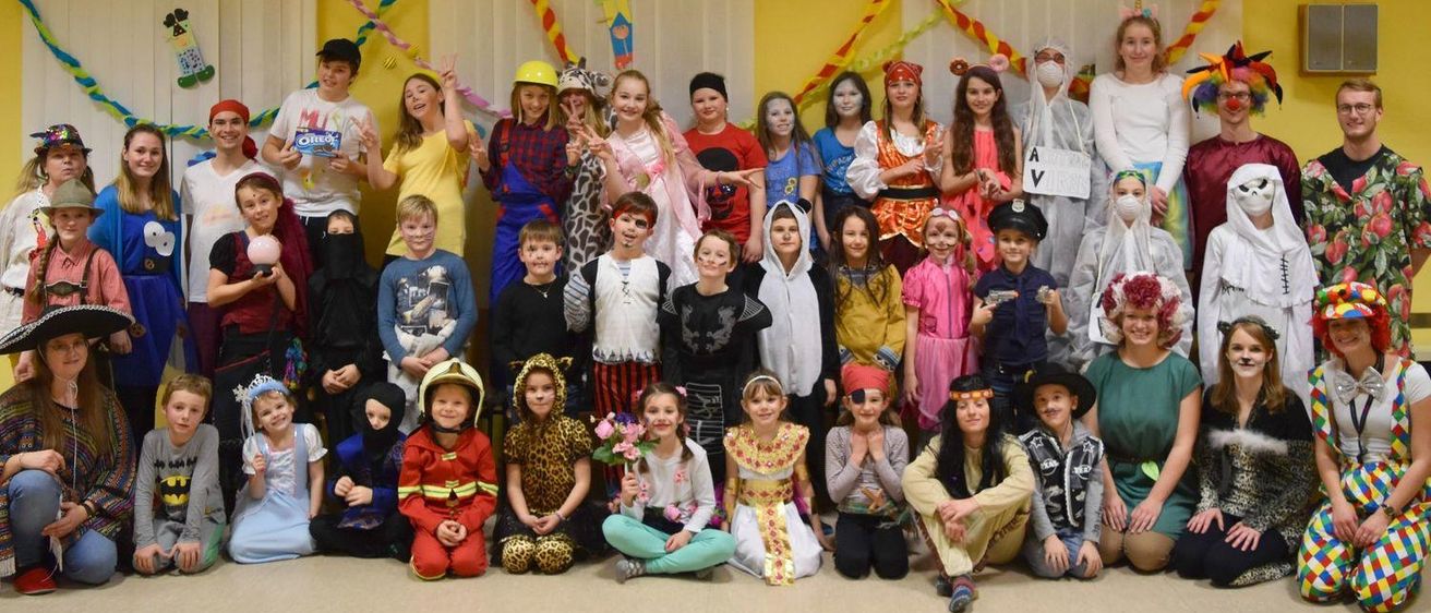 Eine Gruppe von Kindern und Erwachsenen in verschiedenen Kostümen posiert für ein Foto in einem Raum mit gelben Wänden und bunten Dekorationen.