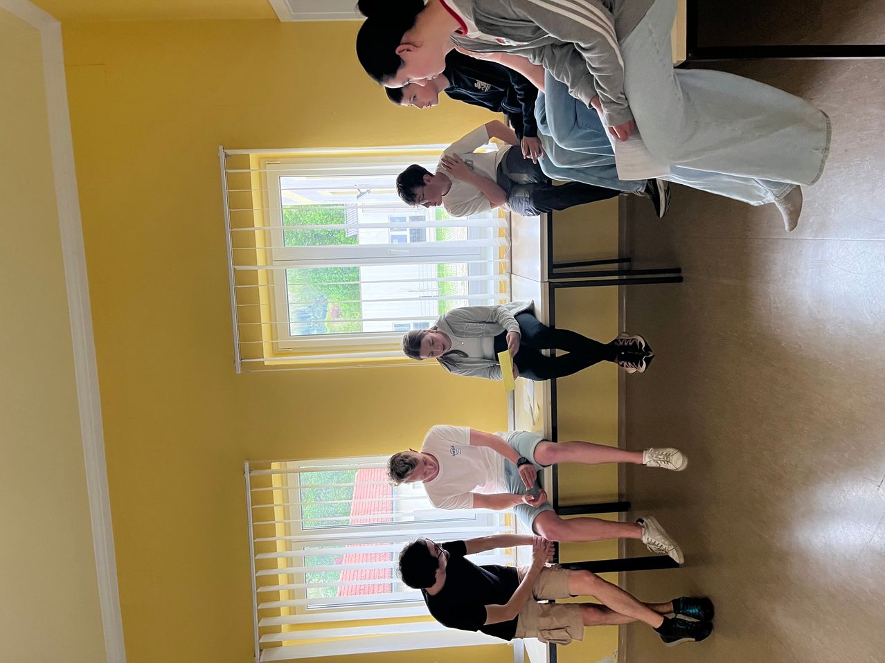 An overhead view of five individuals in a classroom setting, with two individuals standing and three sitting on chairs, one of whom is holding a yellow piece of paper.