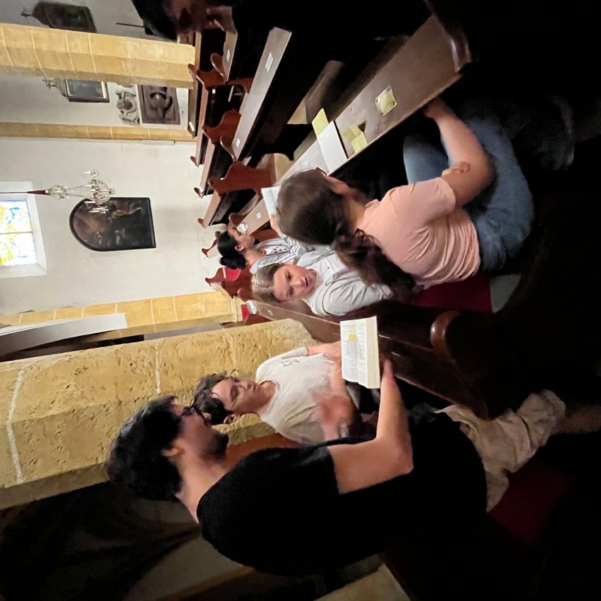 A group of people are sitting in pews inside a church, looking at a book held by one of them. The church has wooden pews, yellow walls, and a stained glass window.