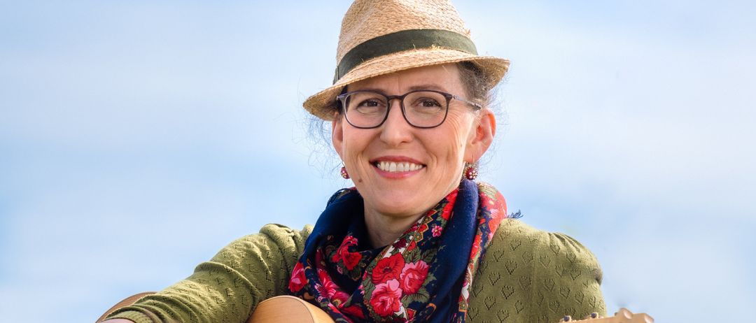 Eine Frau sitzt in einem Grasfeld, lächelt und hält eine Gitarre. Sie trägt einen Hut, eine Brille und eine Schürze. Der Himmel ist blau.