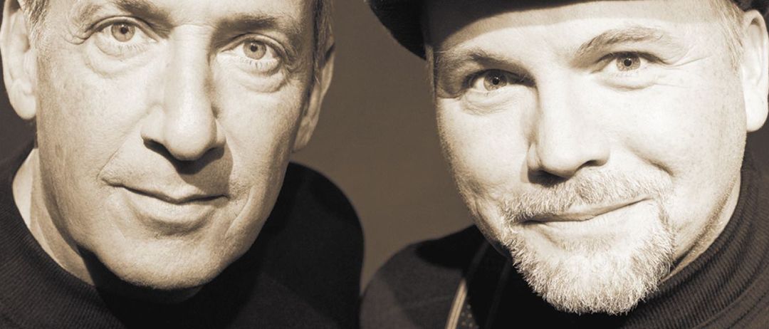 Zwei Männer in einem monochromen Foto, einer mit Bart und Hut, der andere clean-rasiert. Sie schauen direkt in die Kamera mit ernsten Gesichtern.