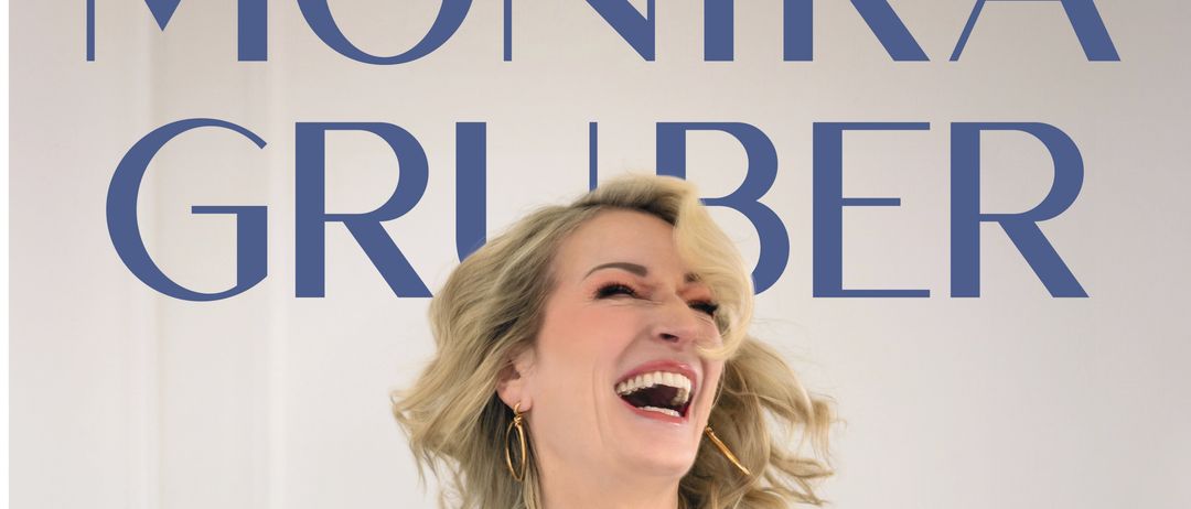 Eine blonde Frau mit fröhlichem Gesichtsausdruck steht vor weißem Hintergrund. Sie trägt einen blauen Nadelstreifenblazer und ein weißes T-Shirt. Der Text 'es huift ja nix' ist in Pink, mit einem Herzsymbol am Ende.