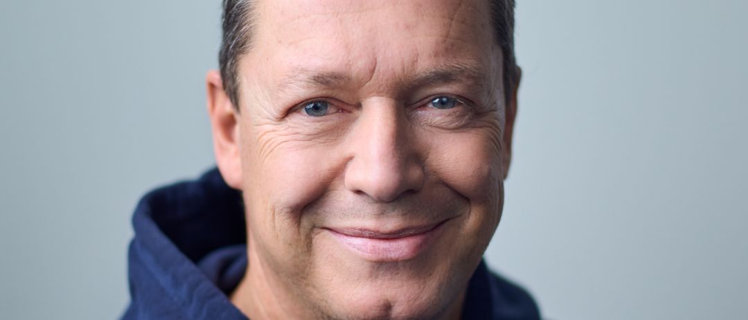 Ein Mann mit grauen Haaren und einer navyblauen Kapuzenjacke lächelt in die Kamera. Er hat leichte Falten um die Augen und eine leichte Stirnrunzel.