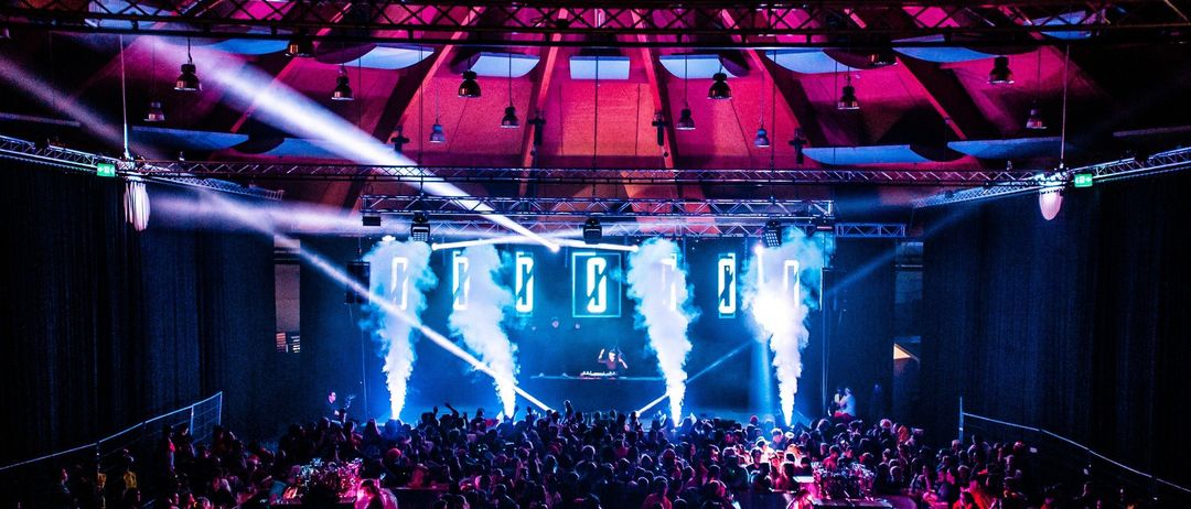 Ein überfüllter Musikclub mit Bühne, bunten Lichtern und Raucheffekten. Die Menschenmenge ist groß, viele Leute genießen die Aufführung. Die Location hat eine hohe Decke mit hängenden Lampen.