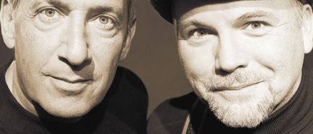 Zwei Männer in Schwarz, einer mit Bart und Hut, der andere mit Schnurrbart, posieren für ein Foto mit ernsten Gesichtern vor einem braunen Hintergrund.
