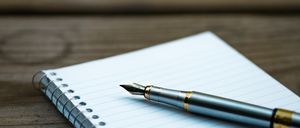 Ein Füllfederhalter liegt auf einem Spiralnotizbuch mit blau kariertem Papier auf einer Holzoberfläche.