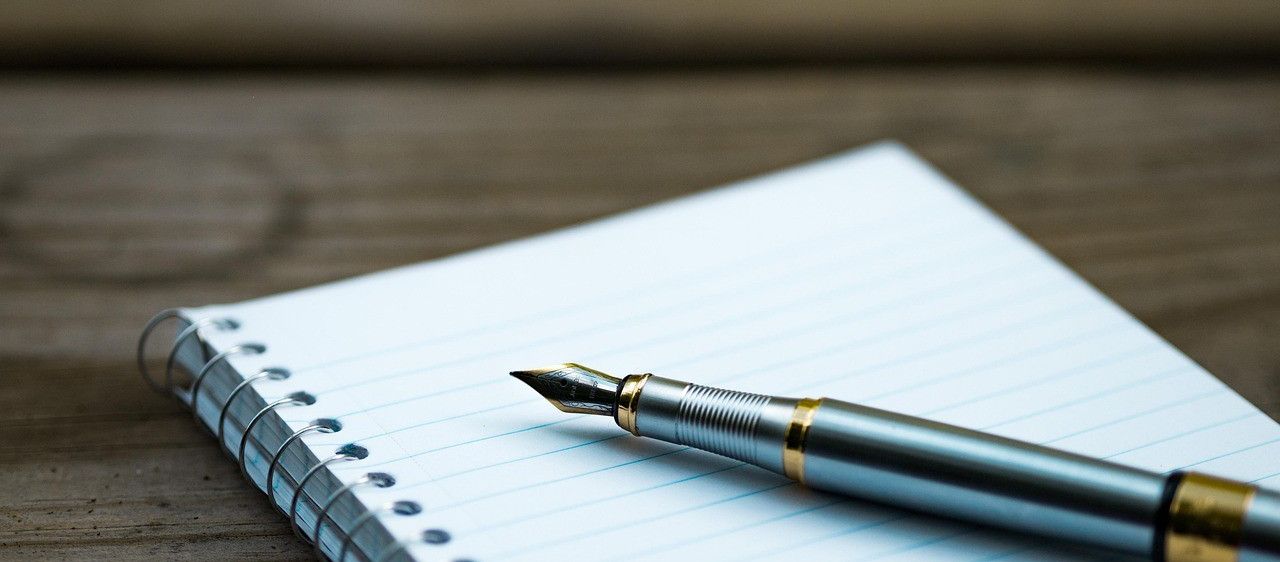 Ein Füllfederhalter liegt auf einem Spiralnotizbuch mit blau kariertem Papier auf einer Holzoberfläche.