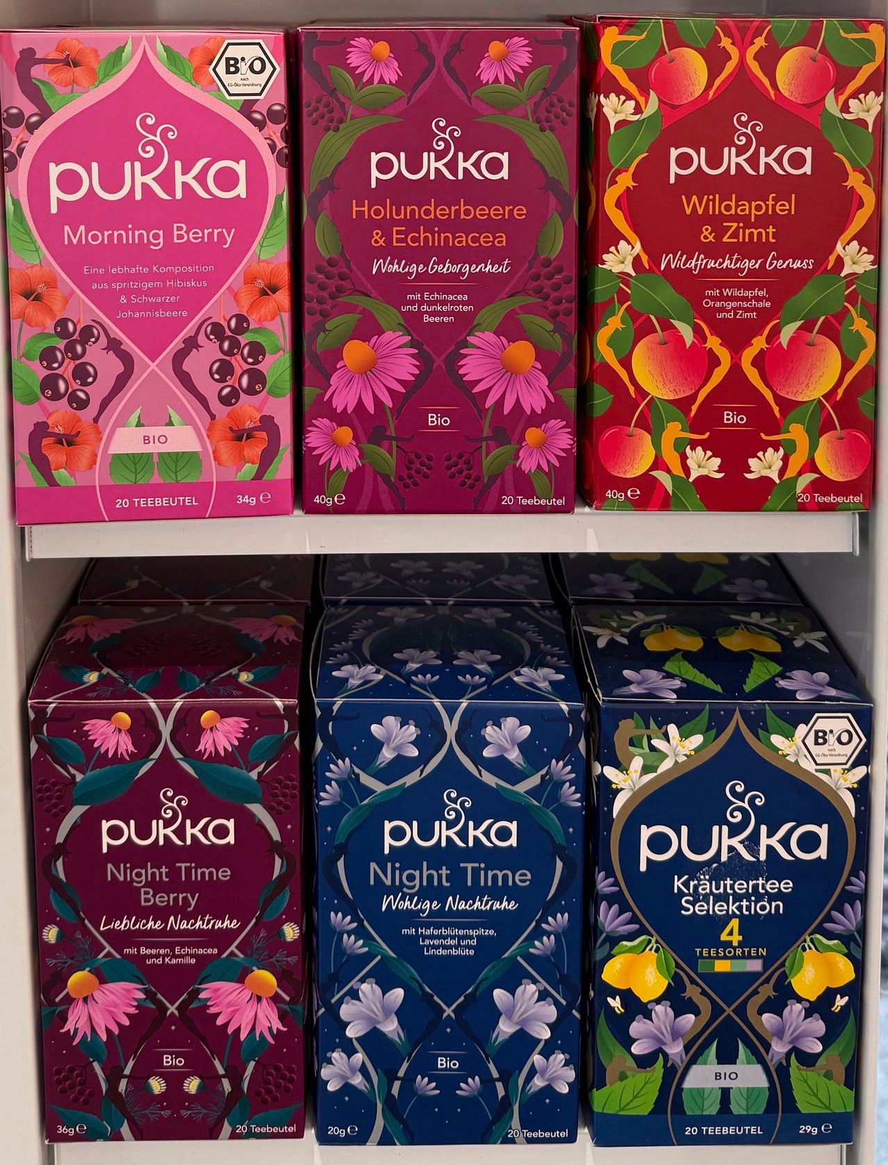 A shelf displays four types of Pukka teas. Two at the top are labeled Morning Berry and Holunderbeere & Echinacea, while the bottom two are labeled Night Time Berry and Kräutertee Selektion.