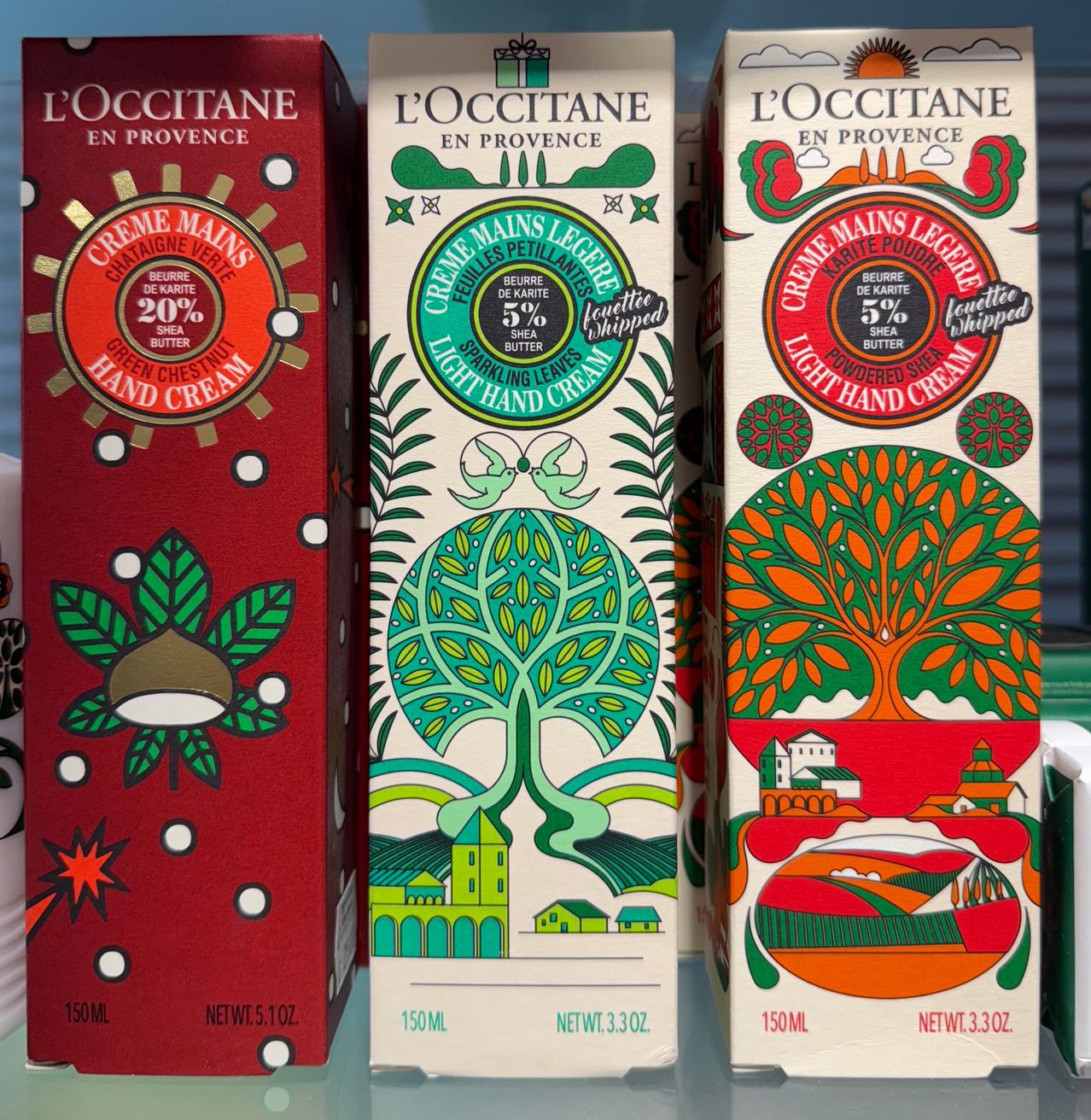 Drei Schachteln L'Occitane Handcremes mit jeweils einem anderen Design und einem Fassungsvermögen von 150ml. Eine Schachtel hat ein rotes Design, eine andere weiß mit grün und die letzte weiß mit rot. Jede Schachtel hat ein kleines Logo auf der Vorderseite.