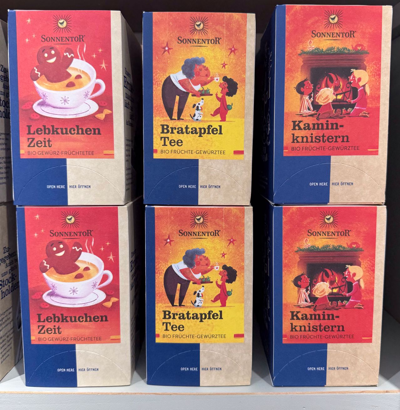 Sechs Schachteln mit Bio-Geschmackstee sind ausgestellt. Sie stammen von der Marke 'Sonnentor'. Die Schachteln sind blau und rot und zeigen verschiedene Illustrationen. Die oberen drei Schachteln sind mit 'Lebkuchen Zeit', 'Bratapfel Tee' und 'Kamin-knist' beschriftet. Die unteren drei Schachteln wiederholen diese Bezeichnungen.