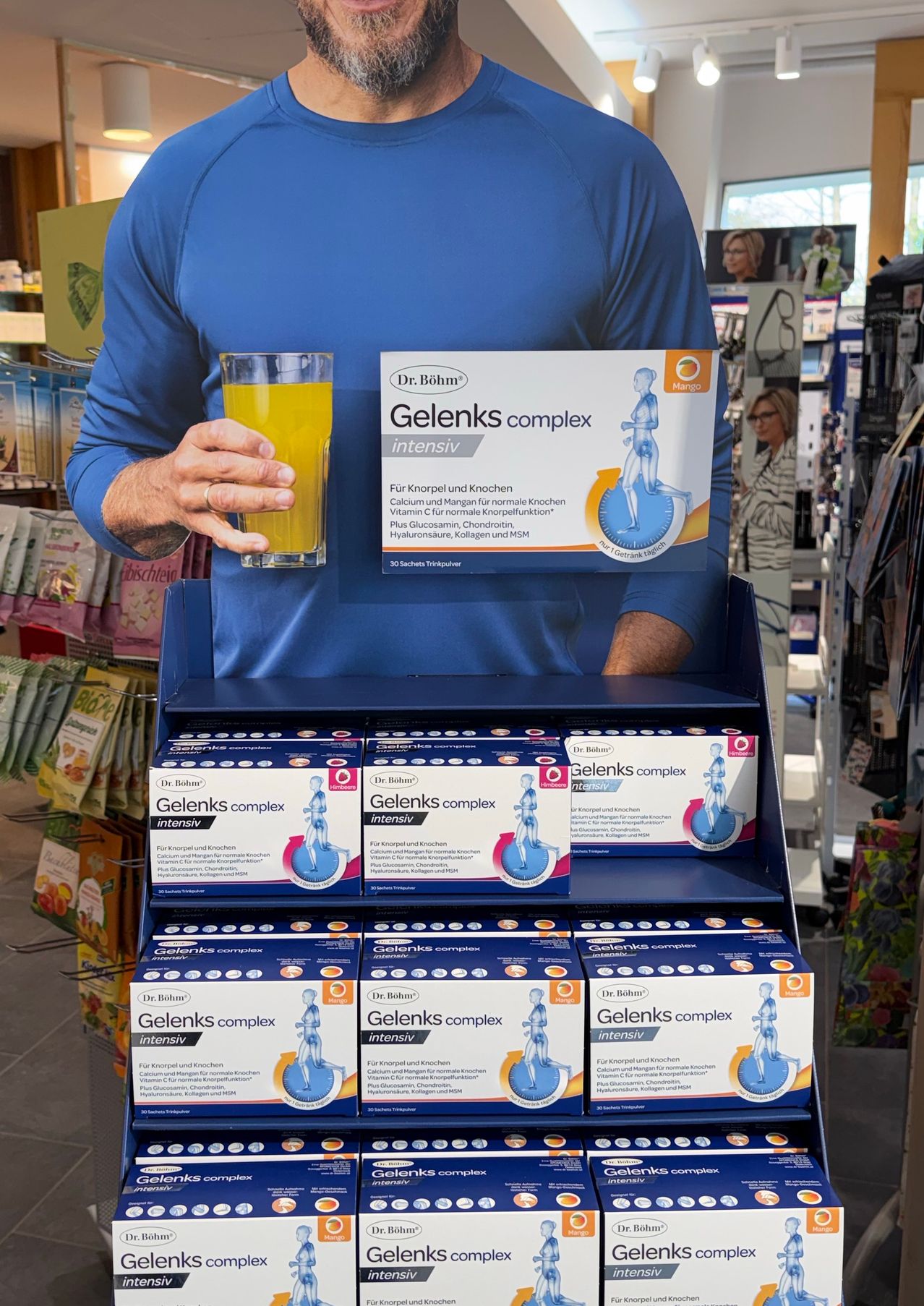 A person holding a glass of juice with boxes of Gelenks complex intensive displayed on a blue shelf.