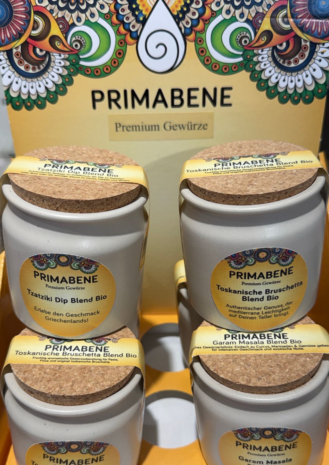 Drei Keramiktöpfe mit Korkendeckeln, beschriftet mit 'Primabened', sind ausgestellt. Jeder Topf hat ein gelbes Etikett mit 'Toskanische Bruschetta Blend Bio' und 'Tzatzik Dip Blend Bio'. Die Töpfe sind auf einer gelben Oberfläche gestapelt.