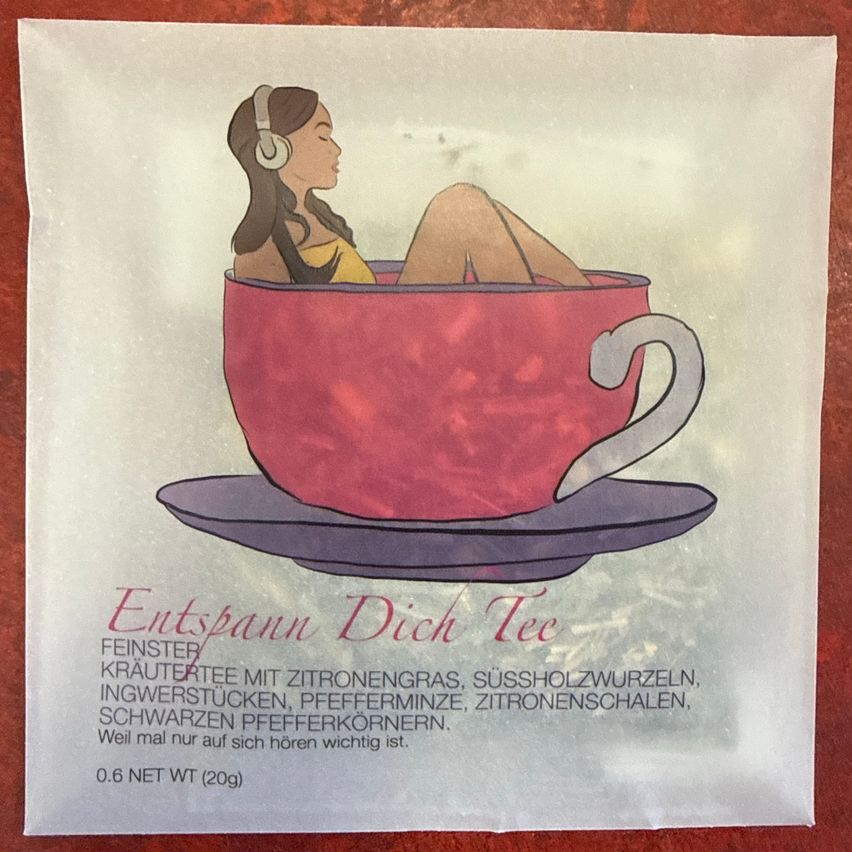 Ein Poster zeigt eine Frau, die in einer roten Teetasse mit Kopfhörern entspannt. Das Poster ist für 'Entspann Dich Tee', ein Kräutertee. Er enthält Zitronengras, Süßholzwurzeln, Ingwerstücke, Pfefferminze, Zitronenschalen, schwarzen Pfefferkörnern. 0,6 NET WT (20g).