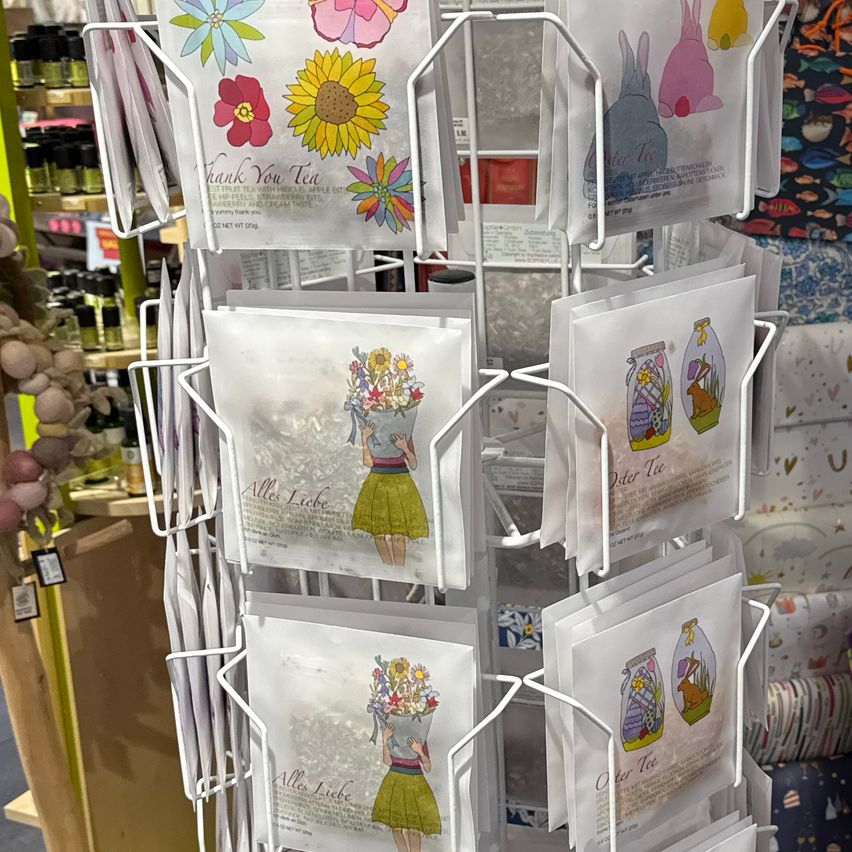 Eine Anzeige von farbenfrohen Postkarten mit Illustrationen wie Blumen und Hasen wird auf einem weißen Drehständer gezeigt. Die Postkarten haben Texte in verschiedenen Sprachen.