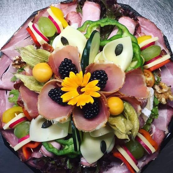 Ein Charcuterie-Brett zeigt eine Blumenanordnung aus Schinken, Käse, Oliven, Trauben und Gemüse, arrangiert auf einem silbernen Teller.